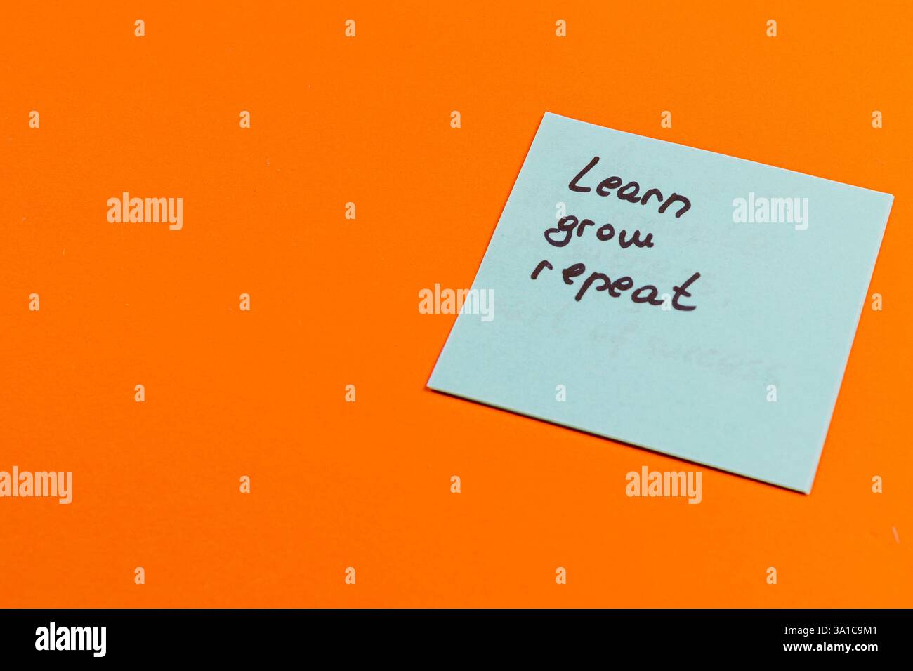 Una nota scritta a mano su un tappetino adesivo azzurro con le parole Learn, Grow e REPEAT si trova su una superficie arancione vibrante, sottolineando un messaggio di contin Foto Stock