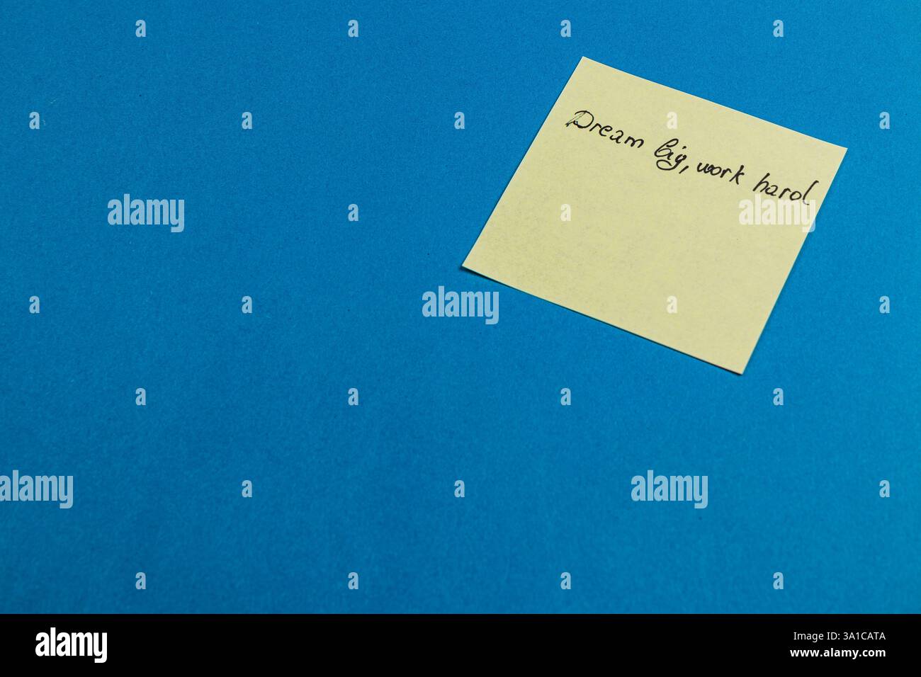 Una nota adesiva con il messaggio Dream Big, Work Hard si appoggia su uno sfondo blu brillante, che serve a ricordare l'ambizione e la dedizione al raggiungimento di g Foto Stock