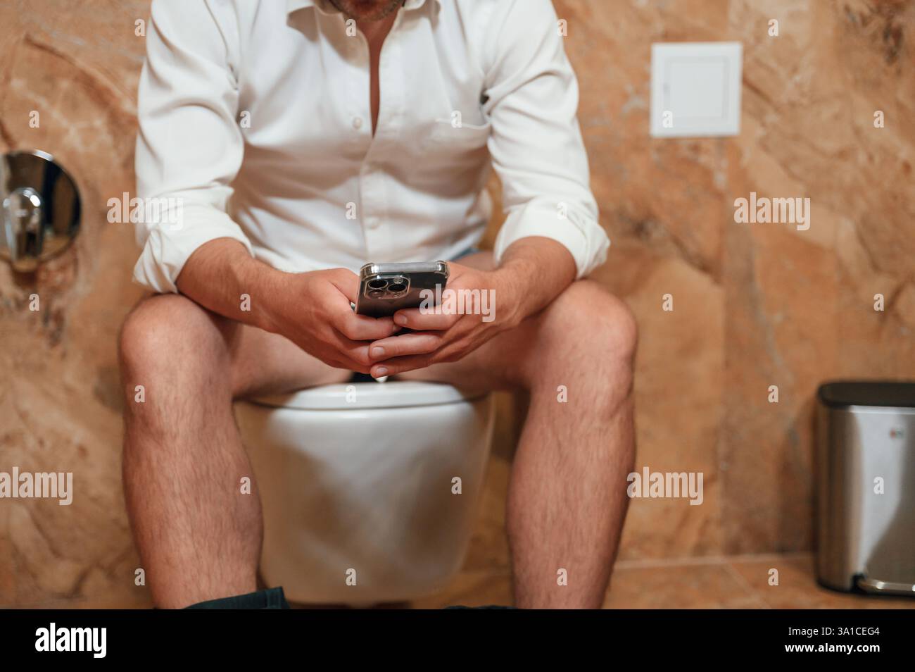 Un uomo con una camicia bianca è seduto sul gabinetto, tenendo uno smartphone tra le mani. Foto Stock