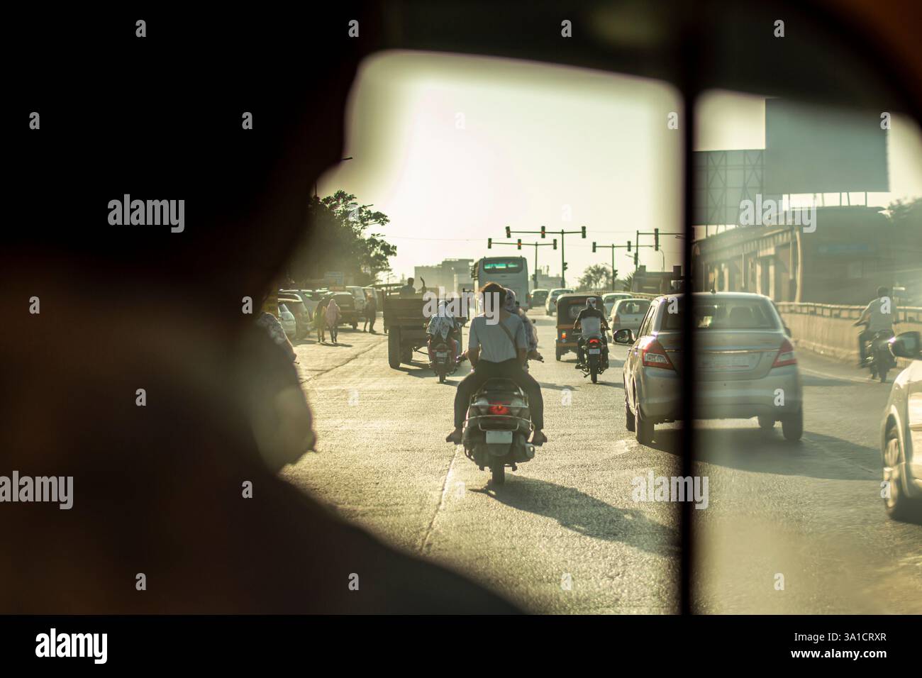Vista dall'interno di un tuk-tuk in India durante l'ora di punta. Motociclette, automobili e pedoni nelle strade caotiche della città, illuminate da un tramonto dorato. Foto Stock