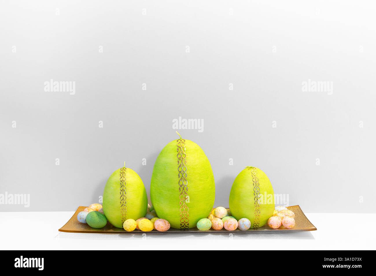 Semplice ed elegante sfondo bianco di Pasqua con candele verdi a forma di uovo di Pasqua sul piatto dorato e piccole uova colorate di quaglia che giacciono intorno. Foto Stock