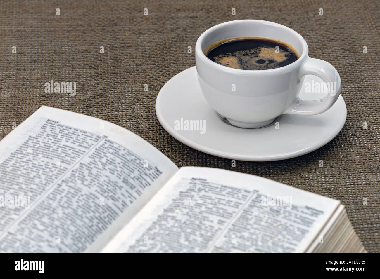 Caffè nero in una tazza bianca su un piattino bianco con un libro e gli indispensabili del mattino sul tavolo. Foto Stock