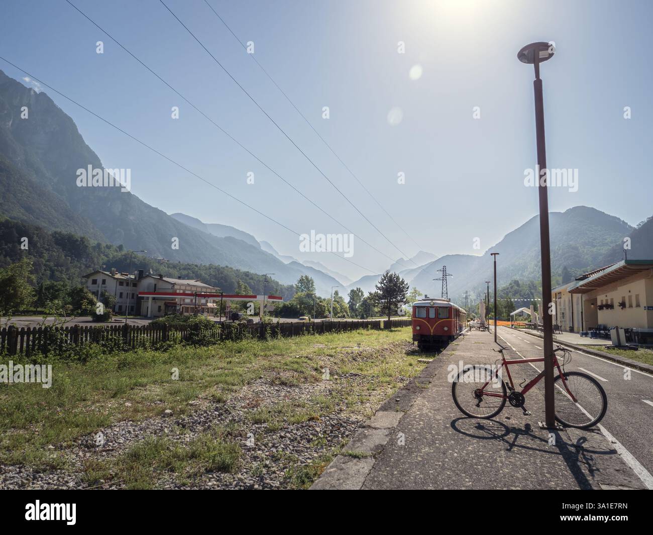 Una bicicletta rossa si appoggia su un lampione in una stazione ferroviaria rurale con un treno rosso d'epoca sullo sfondo. Il paesaggio di montagna e il potere li Foto Stock