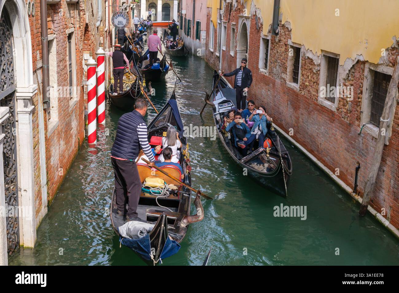 Venezia, Italia - 12 ottobre 2024: Le gondole affollano uno stretto canale mentre i gondolieri in camicie tradizionali a righe navigano lungo il corso d'acqua, trasportando turisti Foto Stock