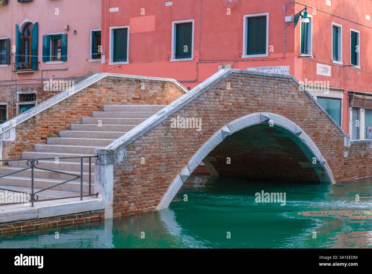 Un tradizionale ponte in mattoni con dettagli in pietra si estende su uno stretto canale, circondato da colorati edifici veneziani Foto Stock