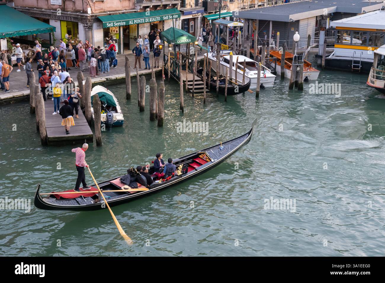 Venezia, Italia - 12 ottobre 2024: Una gondola con passeggeri scivola lungo il canale vicino al ponte di Rialto con turisti nelle vicinanze Foto Stock