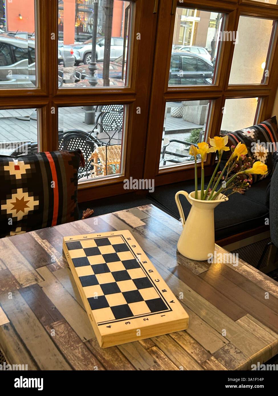 Una scacchiera di legno sul tavolo del ristorante, giocando a gioco cerebrale al coperto con morbidi cuscini, vaso bianco con fiori, finestra, interni dal design elegante Foto Stock