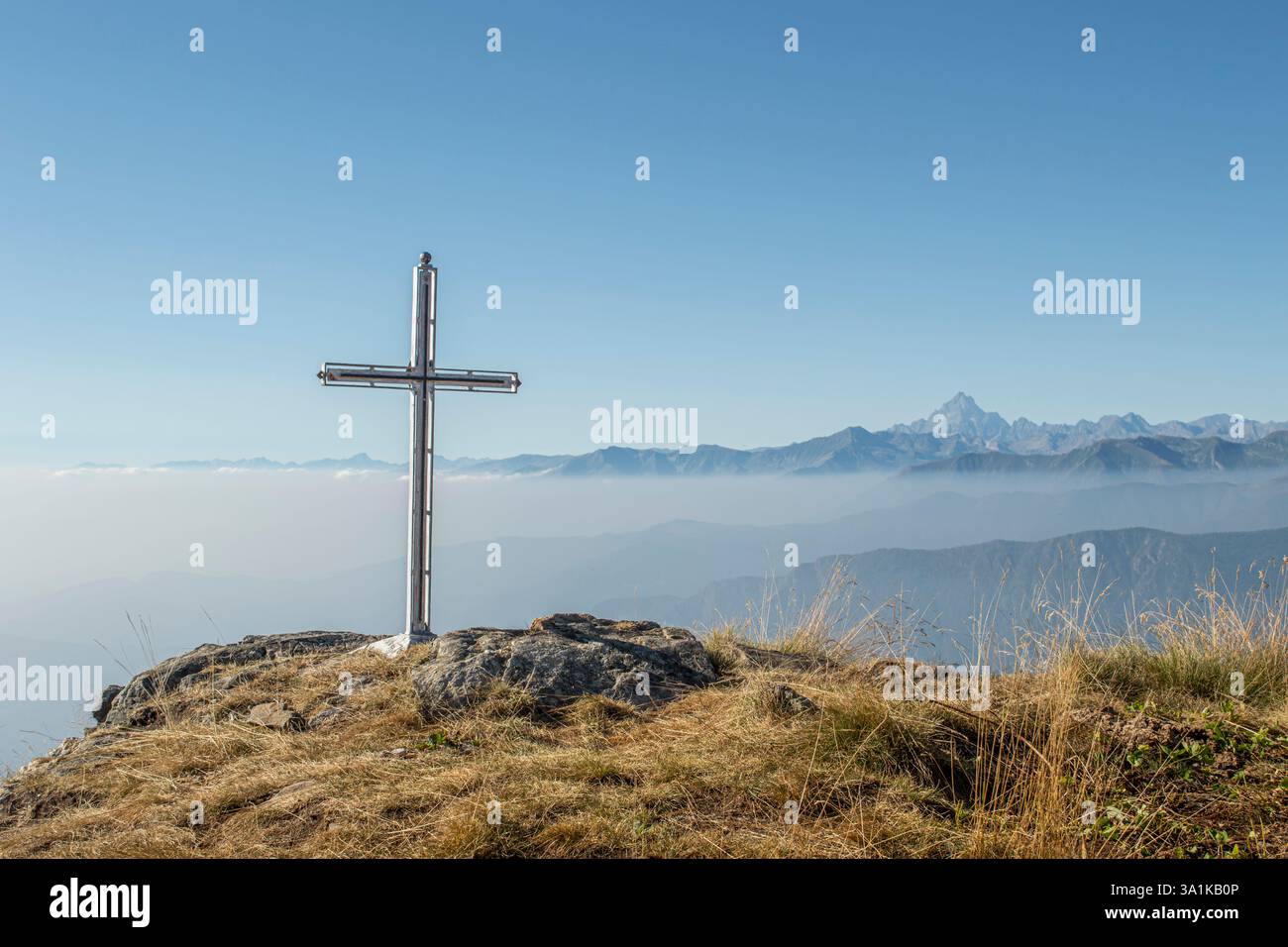 Una vista panoramica delle montagne alpine con una croce in cima in primo piano. La valle nebbiosa sottostante aggiunge profondità e un senso di tranquillità. Foto Stock
