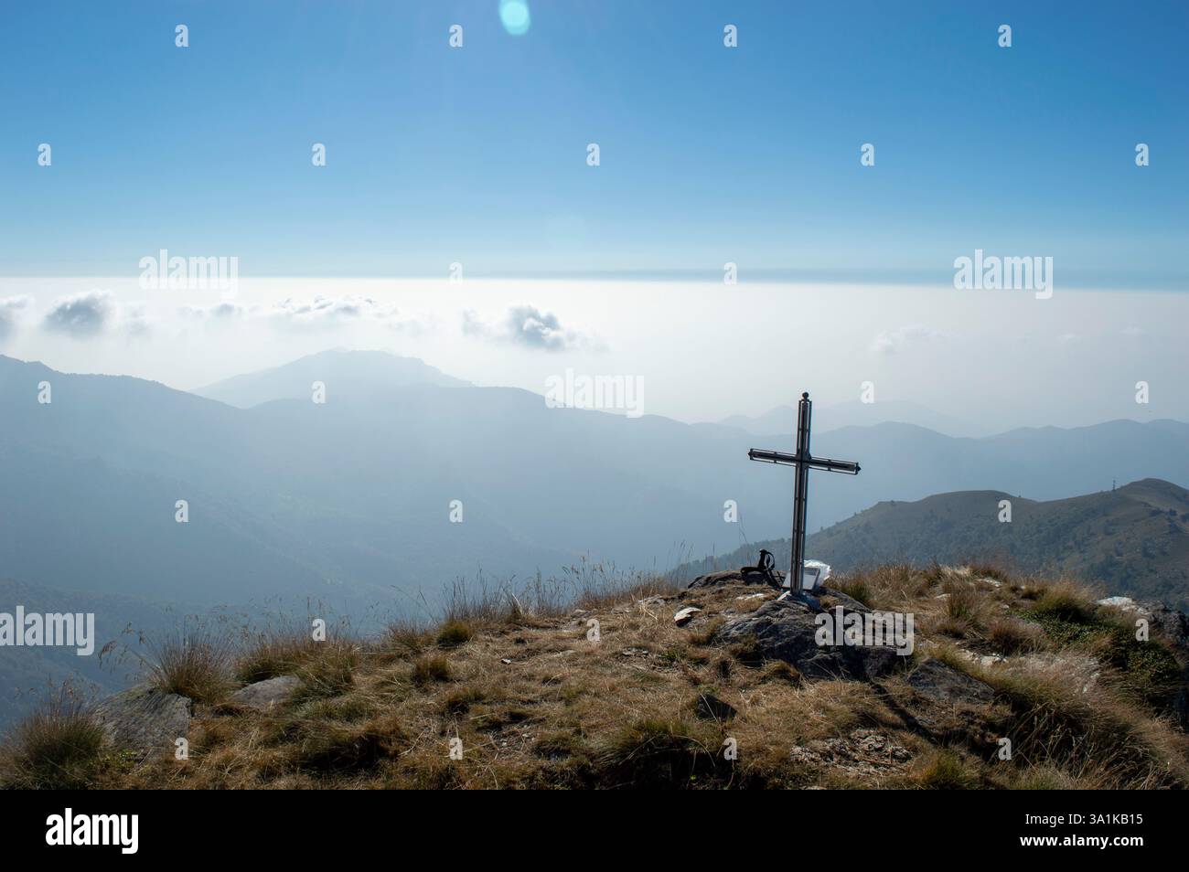 Una vista panoramica delle montagne alpine con una croce in cima in primo piano. La valle nebbiosa sottostante aggiunge profondità e un senso di tranquillità. Foto Stock