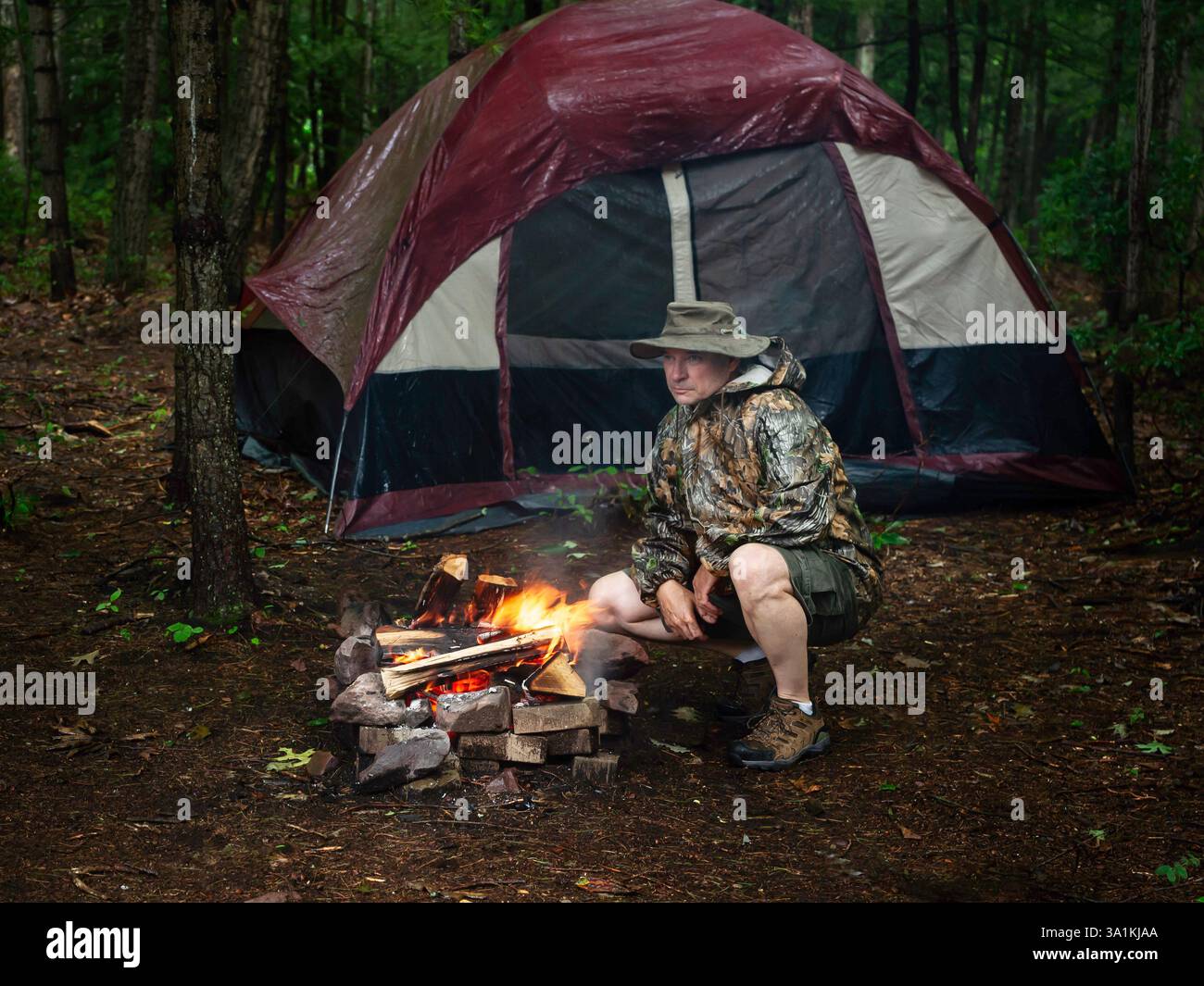 Uomo che si inginocchia vicino al fuoco nella foresta mentre campeggia con la tenda durante il giorno piovoso, indossando pantaloncini e impermeabile Foto Stock