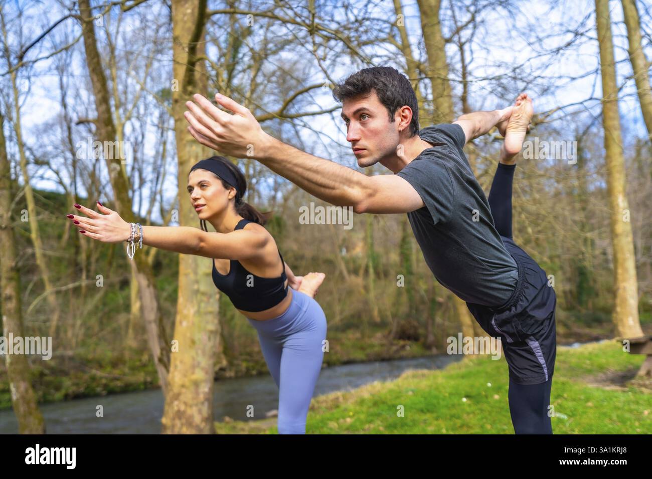 Insegnanti di yoga che eseguono un'asana di bilanciamento vicino a un fiume nella foresta, godendo dei benefici della pratica all'aperto Foto Stock