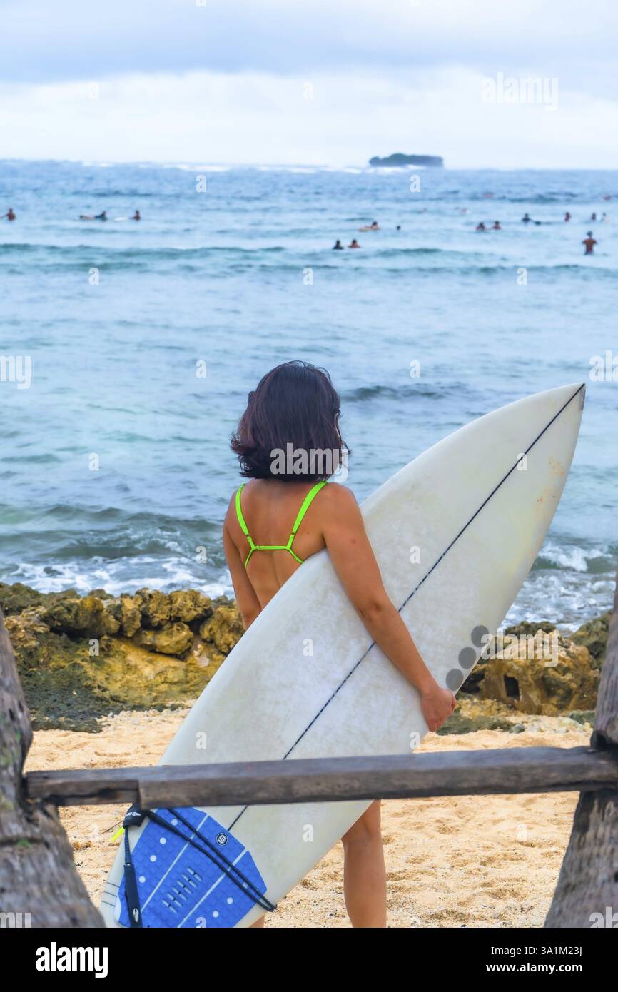 Surfista che trasporta tavole da surf su una spiaggia tropicale nell'isola di Siargao, nelle filippine, osservando altri surfisti nell'oceano Foto Stock