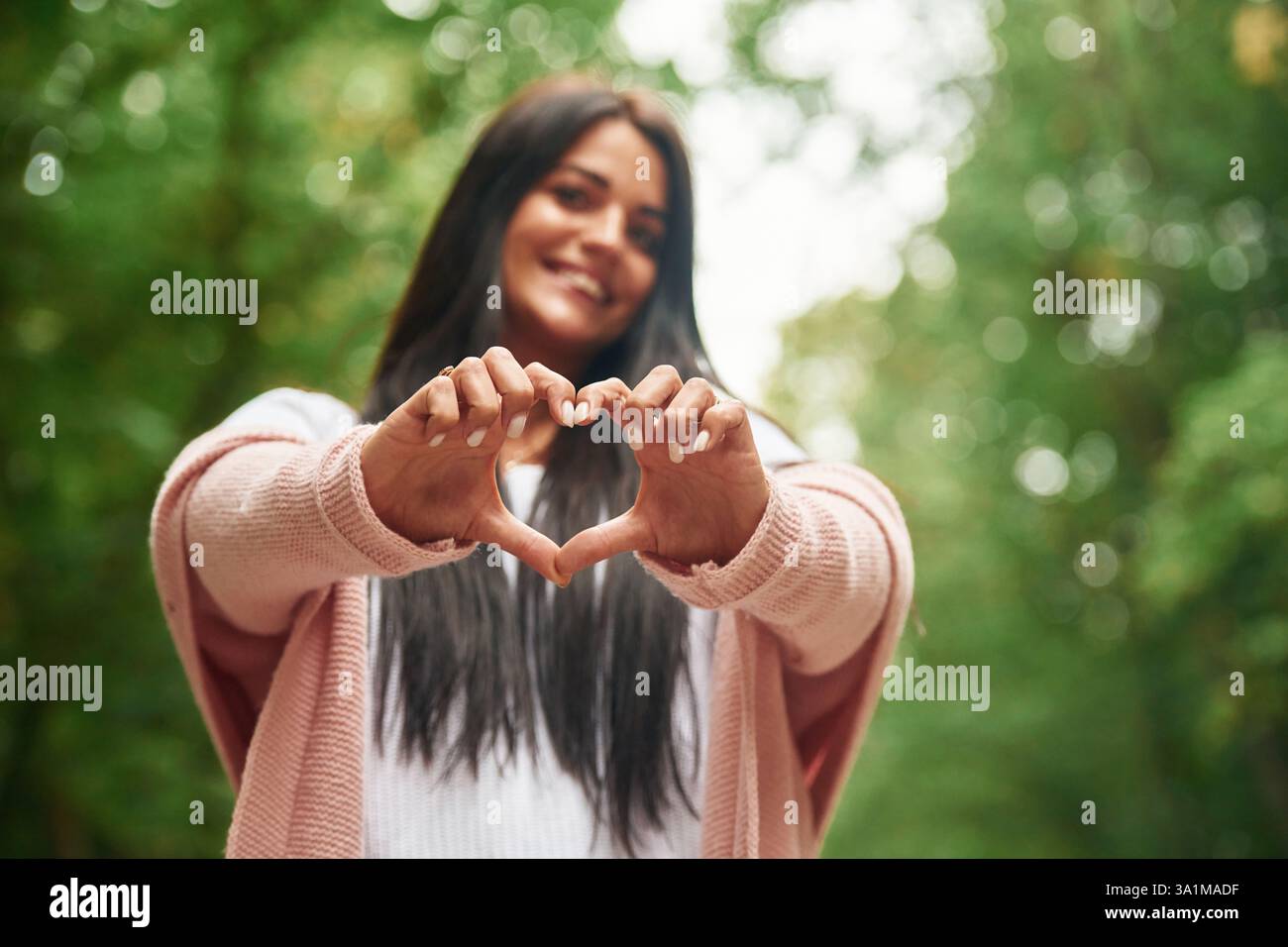 Mostrando un gesto cardiaco con le mani. Allegra bella donna è in piedi all'aperto, foglie verdi sullo sfondo. Foto Stock