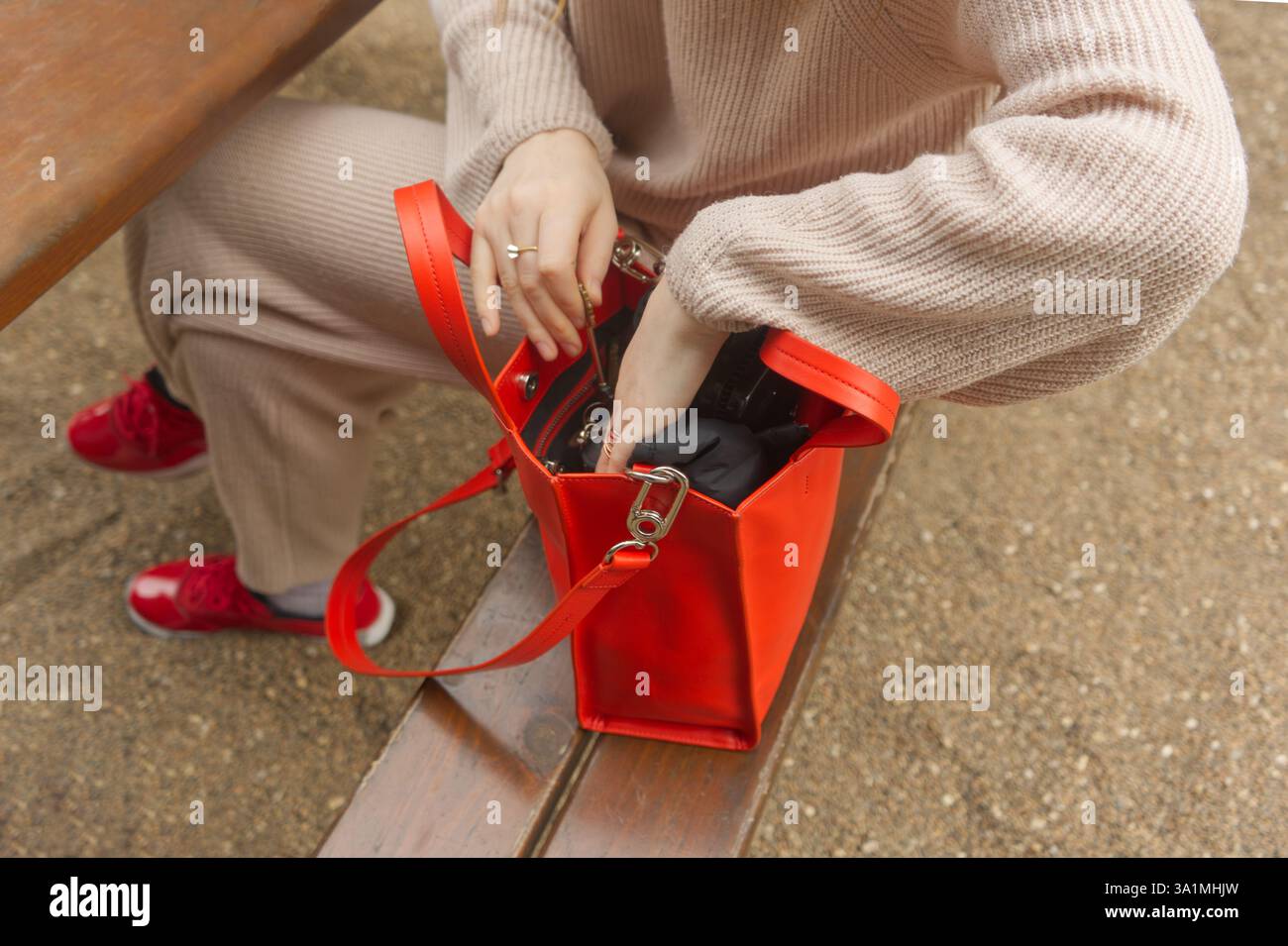 Una persona siede su una panchina, a cercare in una borsa rossa aperta. Il colore rosso intenso contrasta con lo sfondo neutro Foto Stock