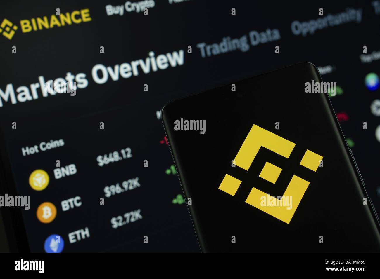 Il logo Binance viene visualizzato sullo schermo di uno smartphone, con il sito Web binance visibile sullo schermo di un computer sullo sfondo Foto Stock