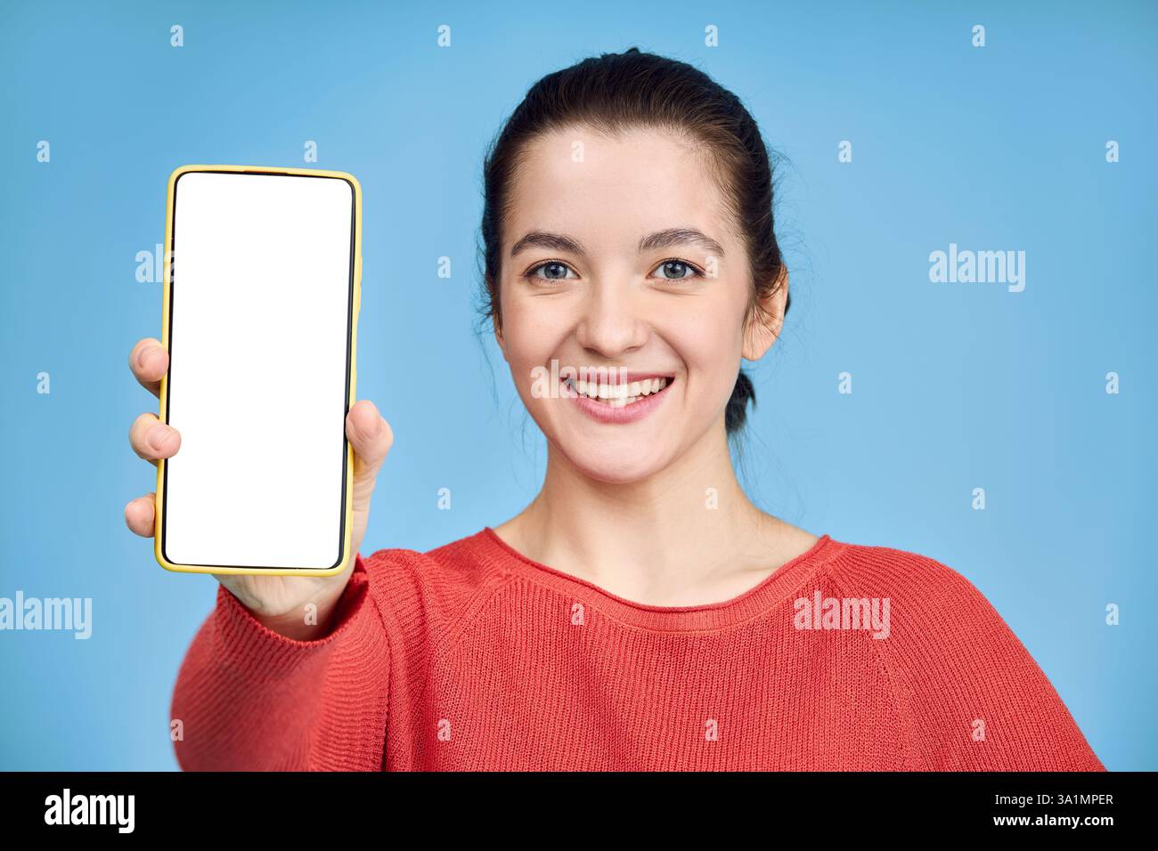 La giovane donna allegra con maglione arancione su sfondo blu mostra un display vuoto del telefono cellulare. Foto Stock
