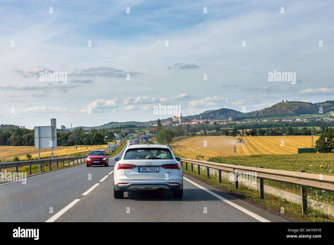 Auto che guidano su strada vicino a Mikulov, Repubblica Ceca, con campi gialli, colline e lo skyline storico della città sotto un cielo estivo parzialmente nuvoloso Foto Stock