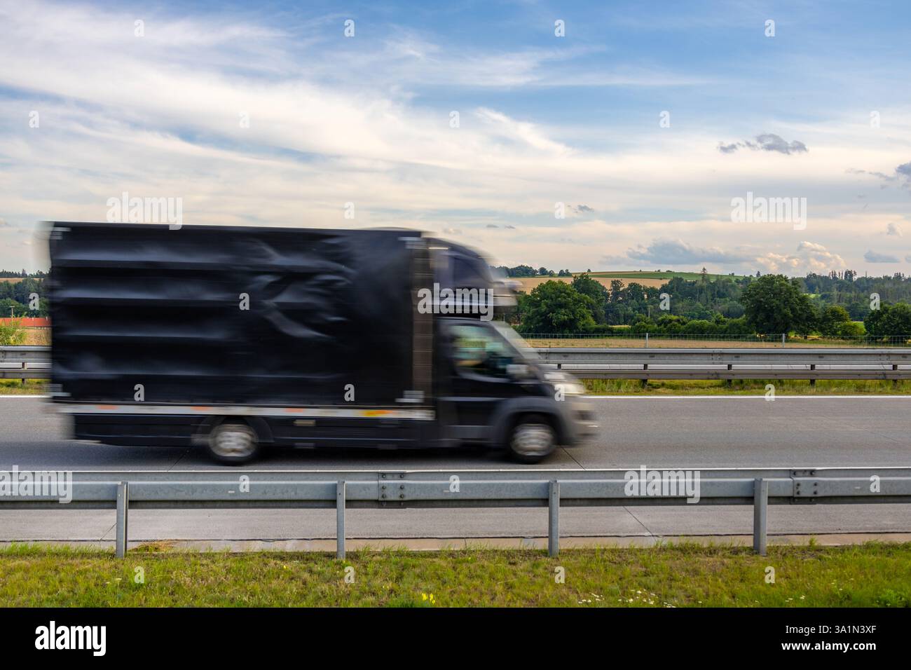 Camion per consegne nero che viaggia in autostrada attraverso la campagna ceca con campi verdi, alberi e un cielo estivo nuvoloso sullo sfondo Foto Stock