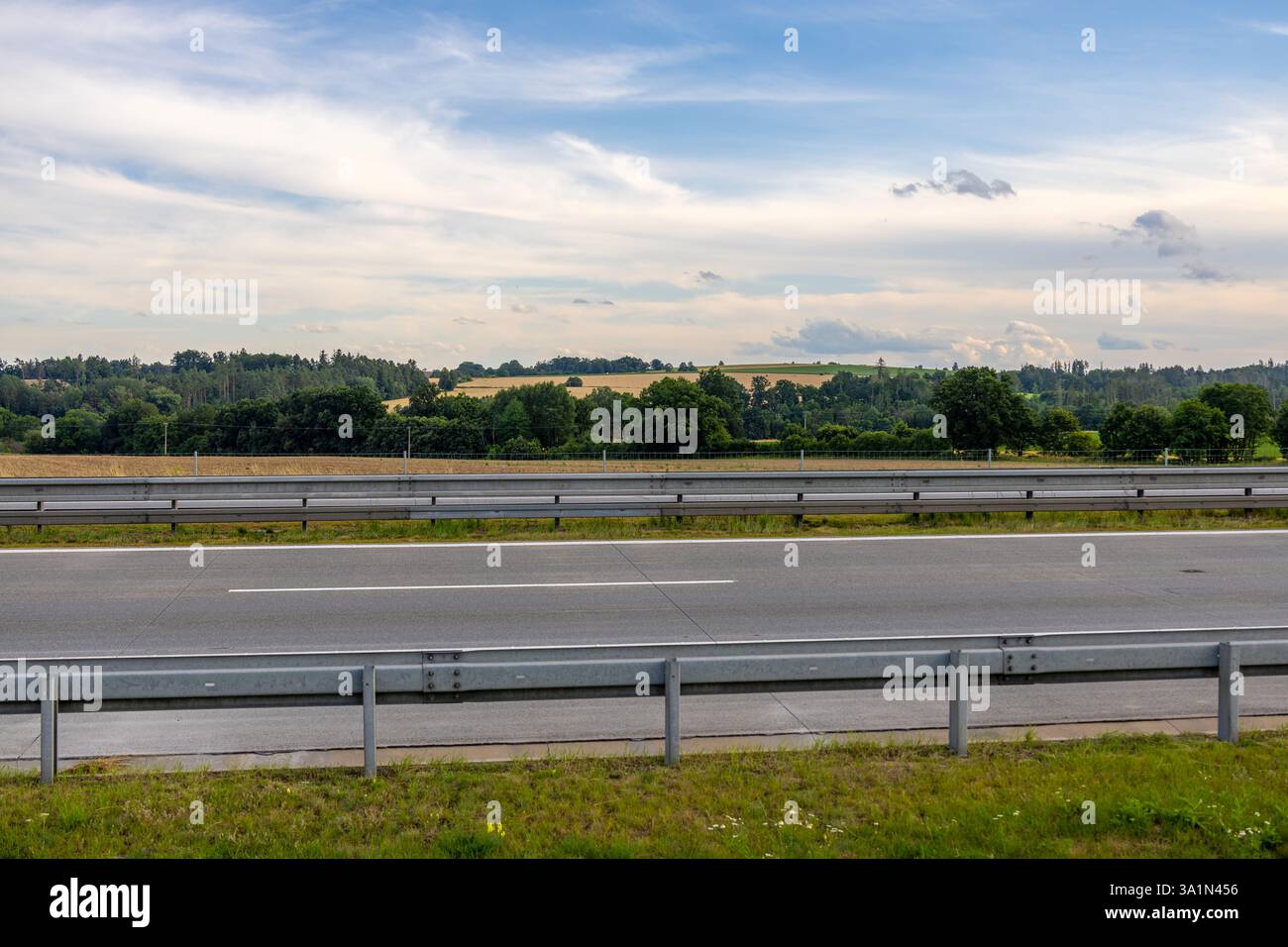Autostrada vuota attraverso la campagna ceca con distanti campi gialli, alberi verdi e un cielo estivo blu nuvoloso Foto Stock