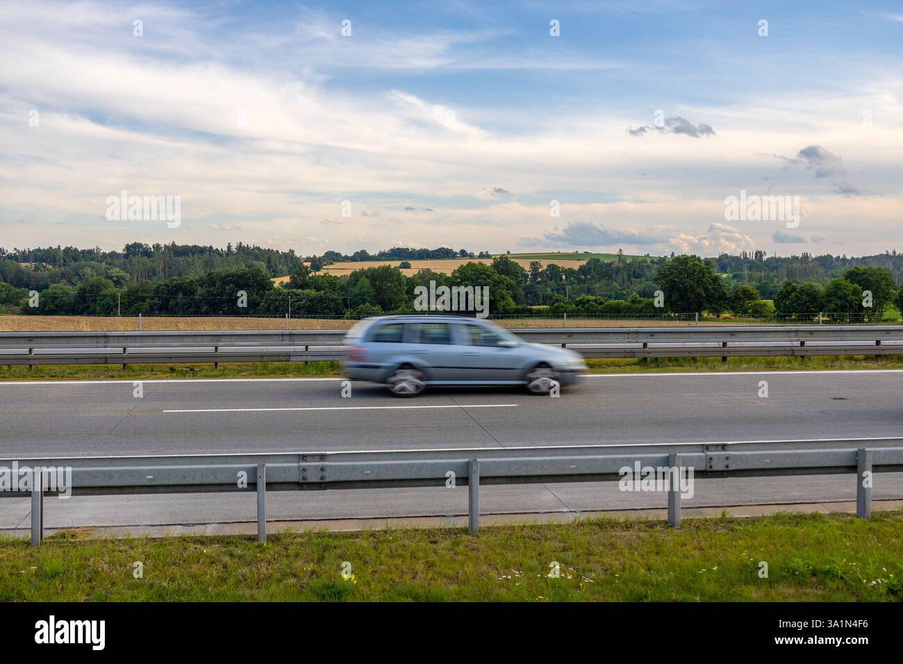 La tenuta Silver Škoda Octavia guida velocemente sull'autostrada ceca passando davanti a campi dorati, alberi verdi e colline boscose sotto un cielo estivo nuvoloso Foto Stock