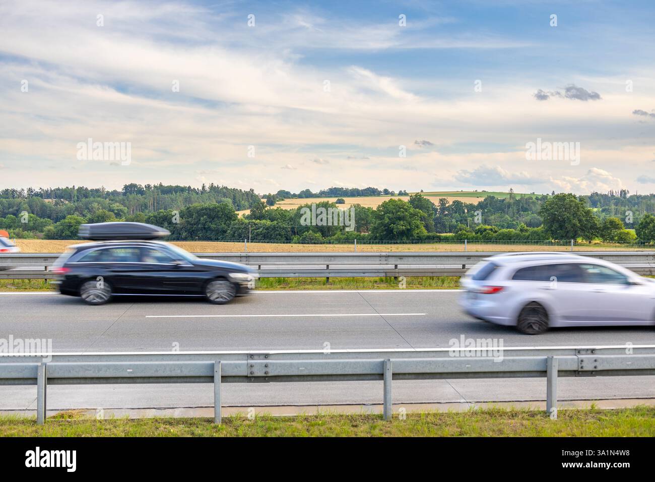 Due auto in movimento sull'autostrada ceca, una nera con box da tetto e una tenuta d'argento, passando per terreni agricoli e alberi sotto il cielo estivo nuvoloso Foto Stock