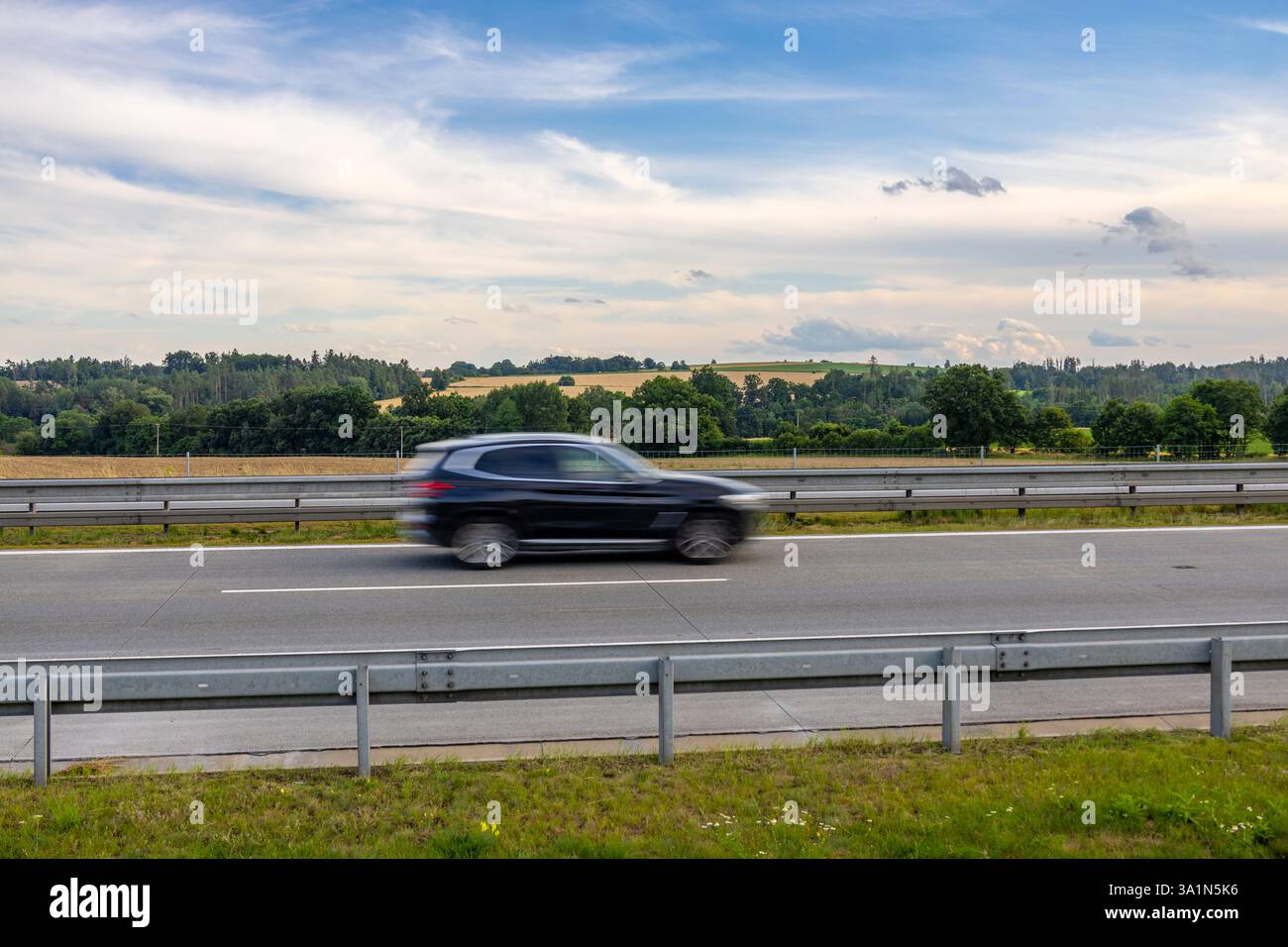 SUV nero che guida velocemente sull'autostrada ceca con campi verdi e dorati, colline boscose e nuvoloso cielo estivo sullo sfondo Foto Stock
