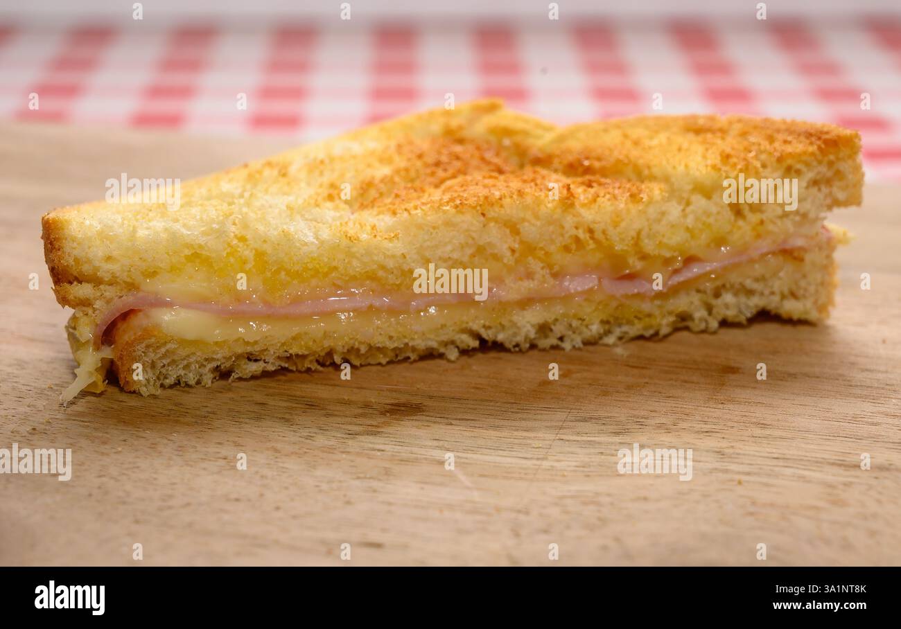 Un panino appena tostato con strati di formaggio e prosciutto, seduto su una superficie di legno con una tovaglia a scacchi rossa e bianca sullo sfondo Foto Stock
