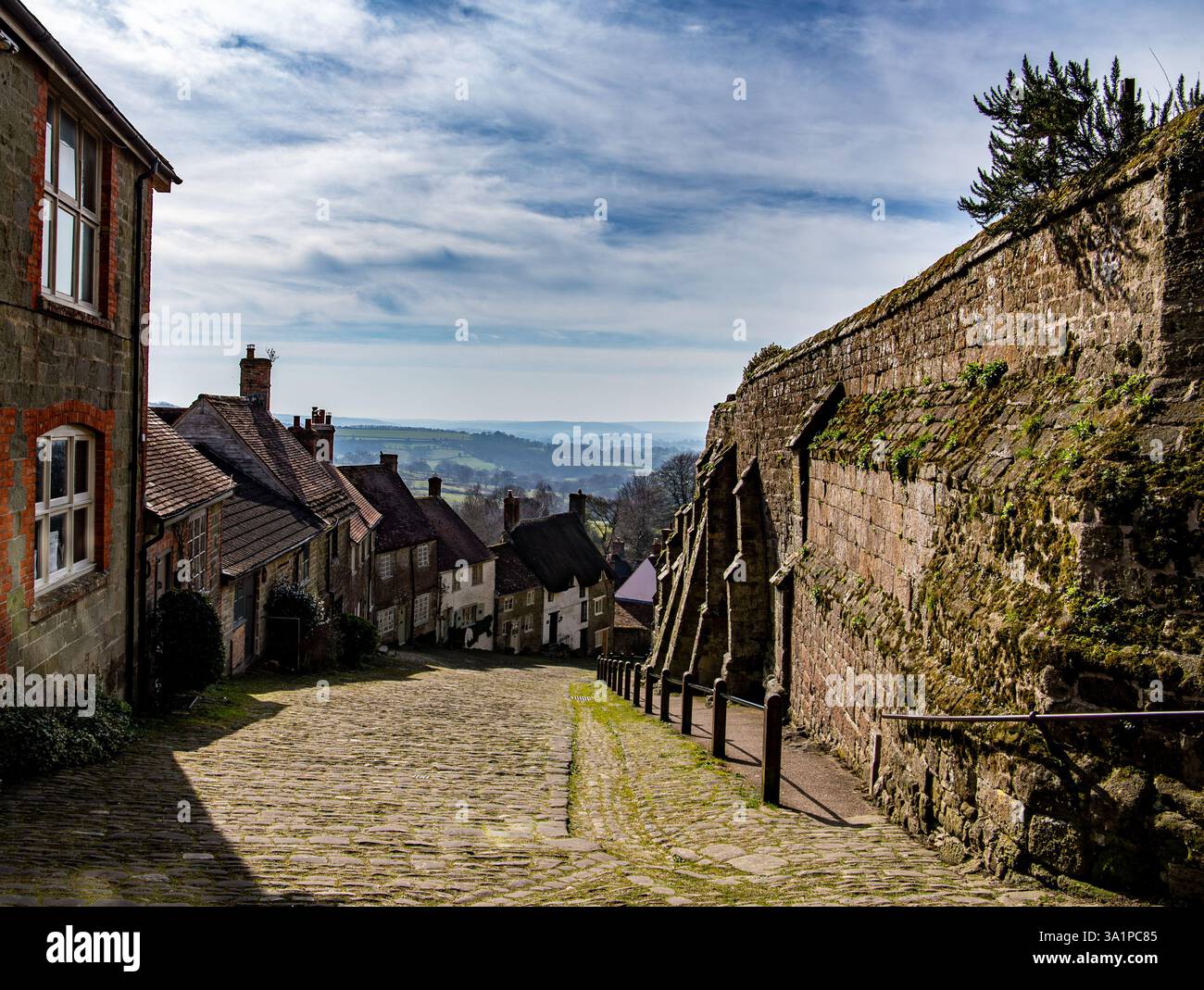 Strada acciottolata fiancheggiata da edifici storici in pietra sotto un cielo parzialmente nuvoloso, con una vista panoramica e distante del Gold Hill Shaftesbury Dorset Foto Stock