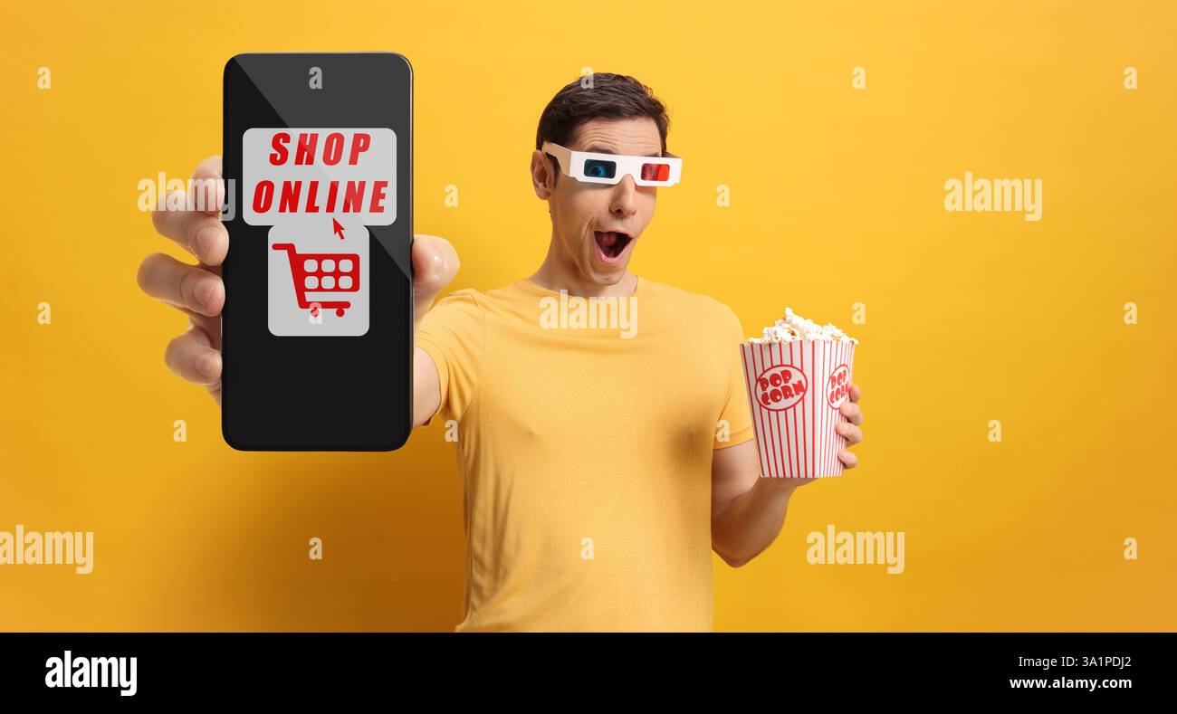 Uomo eccitato con una scatola di popcorn e occhiali da cinema 3D che mostra uno smartphone con negozio di testo online su sfondo giallo Foto Stock