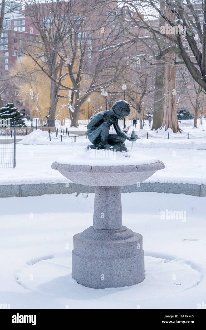 Boston, ma, Stati Uniti - 11 febbraio 2025 - Statua di un bambino seduto che tiene in piedi e fuori con un uccello seduto sulle palme coperte di neve invernale Foto Stock