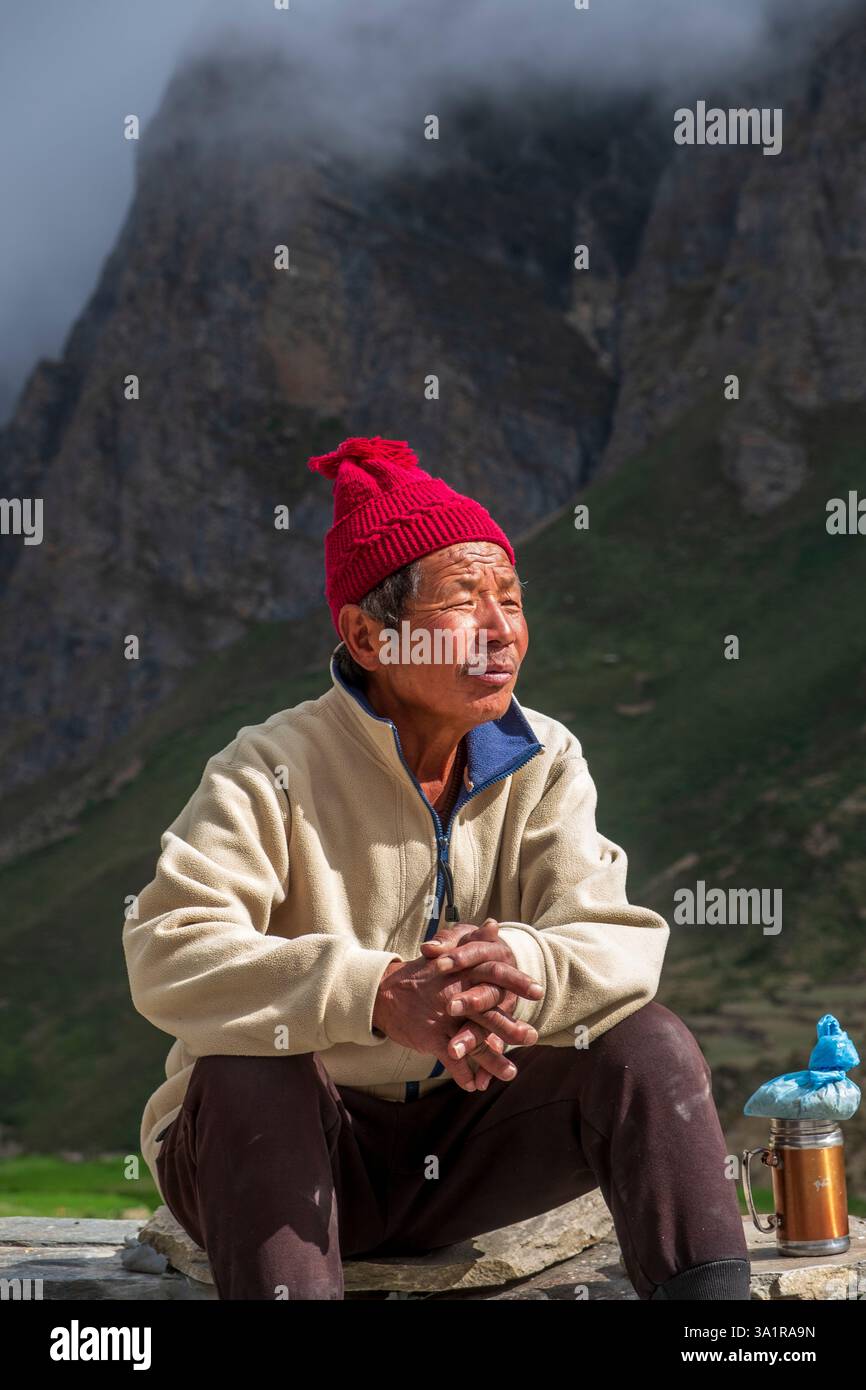Un uomo anziano con un berretto rosso siede pacificamente nel villaggio di Nar in Nepal, circondato da montagne nebbiose. Foto Stock