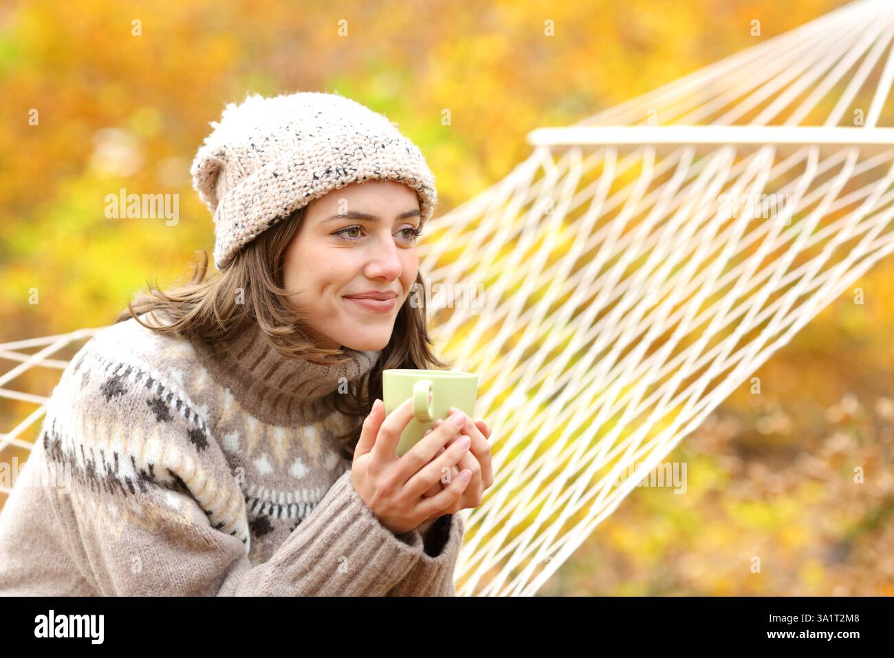 Donna soddisfatta in inverno bevendo caffè guardando lontano seduto su un'amaca in natura Foto Stock