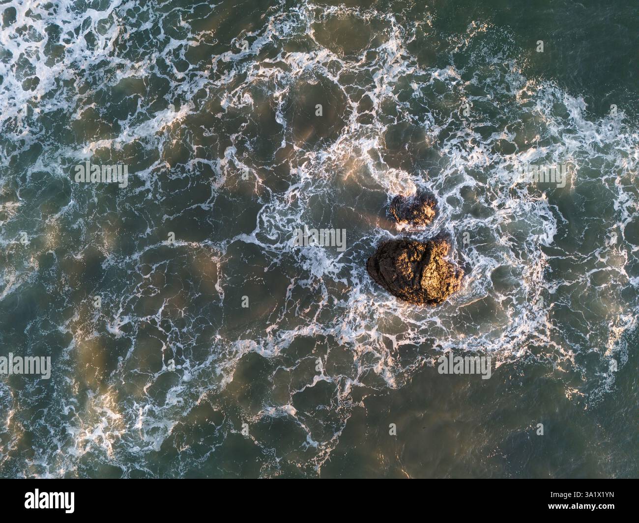 L'affioramento roccioso si erge solitario mentre le onde si infrangono intorno ad esso durante il vibrante tramonto, creando una splendida vista costiera. Foto Stock