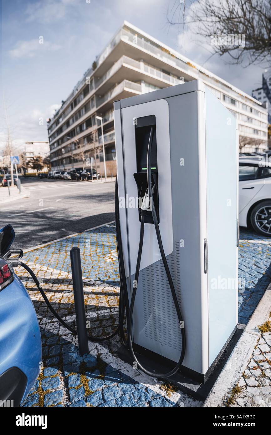 Scatto grandangolare verticale di una moderna stazione di ricarica per veicoli elettrici in un ambiente urbano. Un EV blu è collegato, con i cavi collegati a un elegante Foto Stock