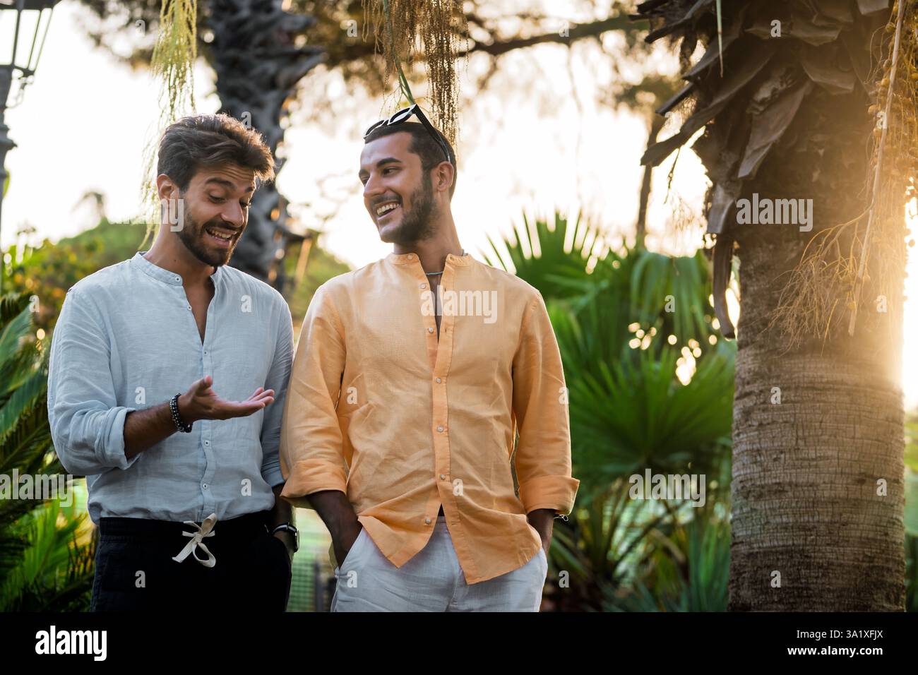 Due giovani uomini alla moda che fanno una conversazione allegra all'aperto in un parco tropicale al tramonto, godendosi un momento informale di amicizia e connessione. Foto Stock Due giovani uomini alla moda che fanno una conversazione allegra all'aperto in un parco tropicale al tramonto, godendosi un momento informale di amicizia e connessione. Foto Stock