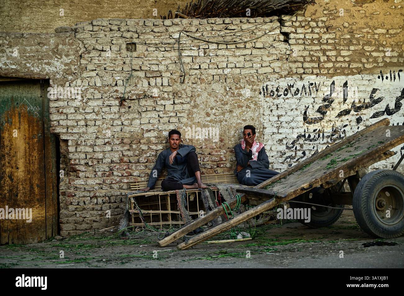 Due agricoltori seduti accanto al loro carro trainato da cavalli fuori da un edificio in mattoni di fango nel villaggio di al-Balyanā, dopo aver appena scaricato il loro raccolto giornaliero Foto Stock