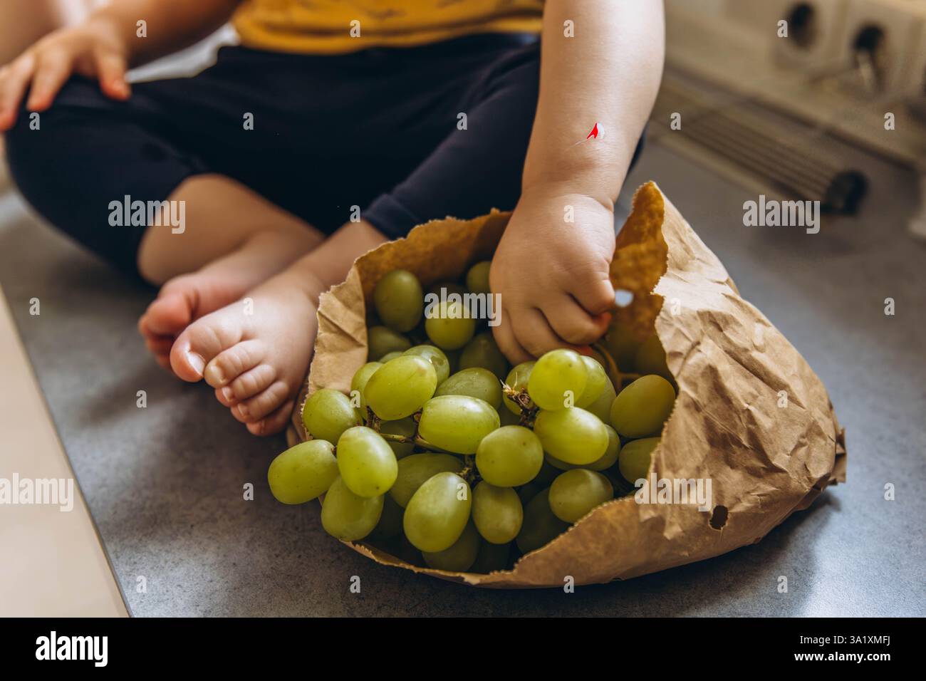 Primo piano dei piedi dei bambini accanto a un sacchetto di carta pieno di uva, per catturare un momento caldo e naturale. Foto di alta qualità Foto Stock