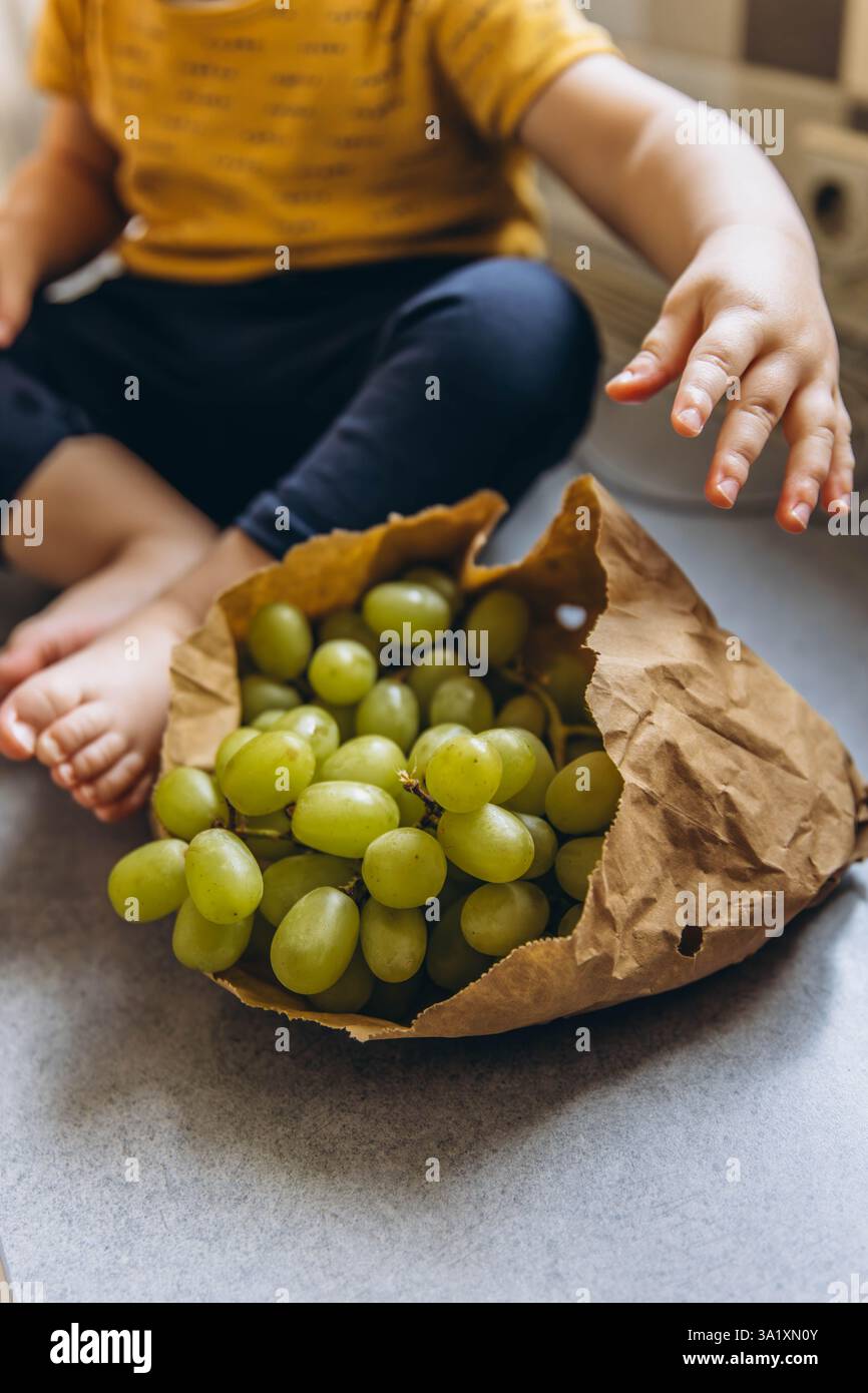 Primo piano dei piedi dei bambini accanto a un sacchetto di carta pieno di uva, per catturare un momento caldo e naturale. Foto di alta qualità Foto Stock