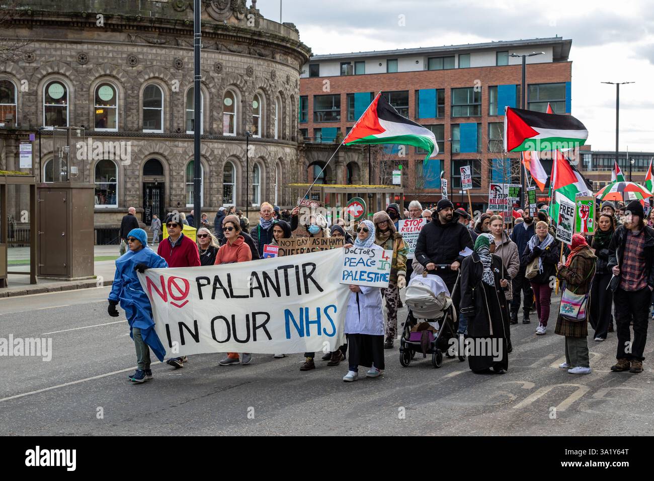 Una folla marcia, tenendo bandiere e cartelli palestinesi, protesta contro il coinvolgimento di Palantir nel NHS. Edifici visibili sullo sfondo. Foto Stock
