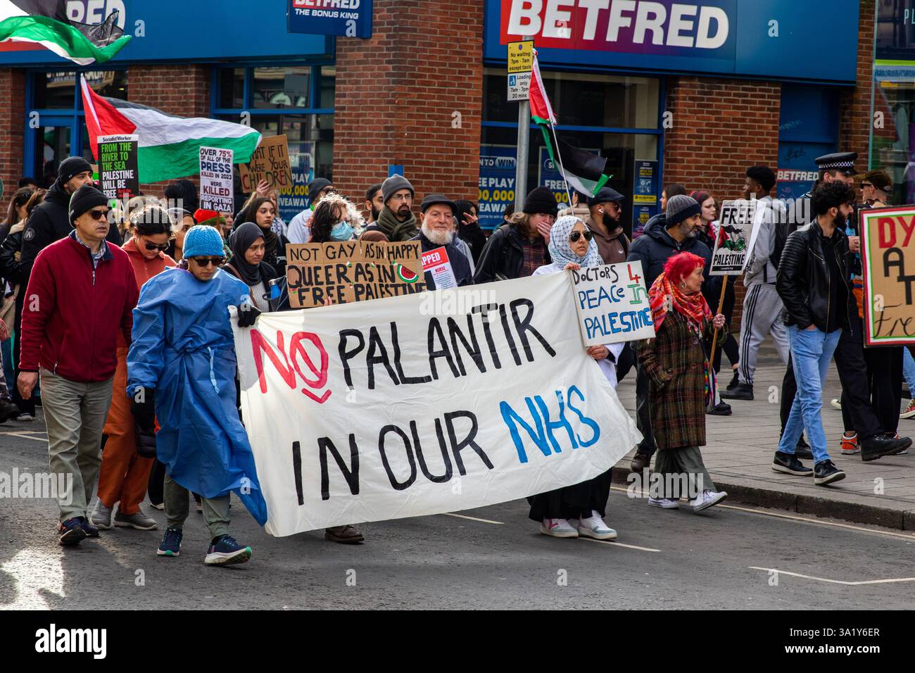 Una marcia di protesta con persone che hanno dei cartelli e delle bandiere palestinesi, protestando contro il coinvolgimento di Palantir nel sistema sanitario nazionale, in un ambiente urbano. Foto Stock