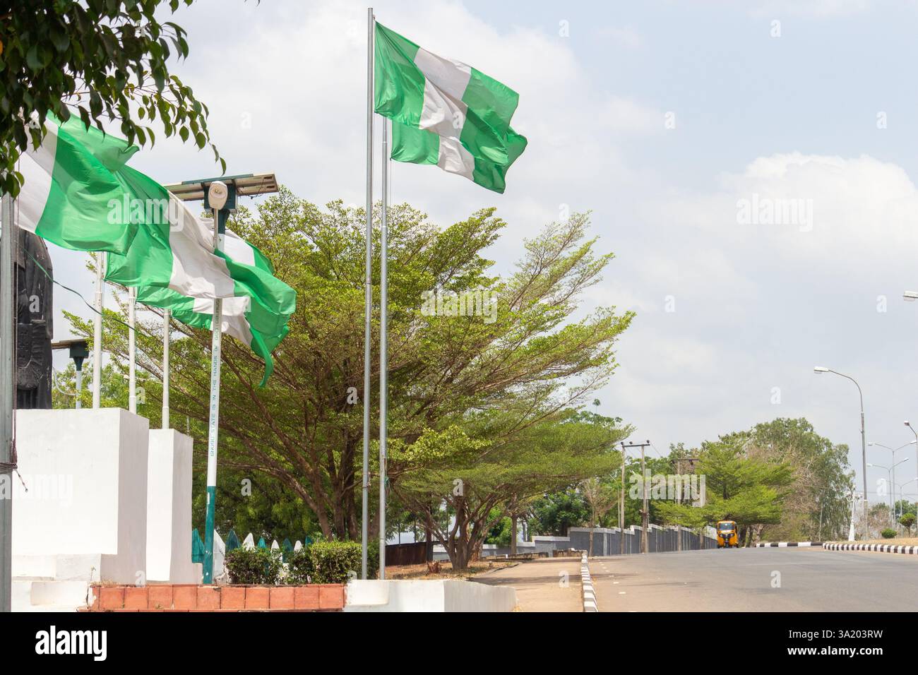 Una strada pulita e ben asfaltata a Enugu, Nigeria, fiancheggiata da bandiere nigeriane e lampioni sotto un cielo parzialmente nuvoloso. Marzo 2025 Foto Stock