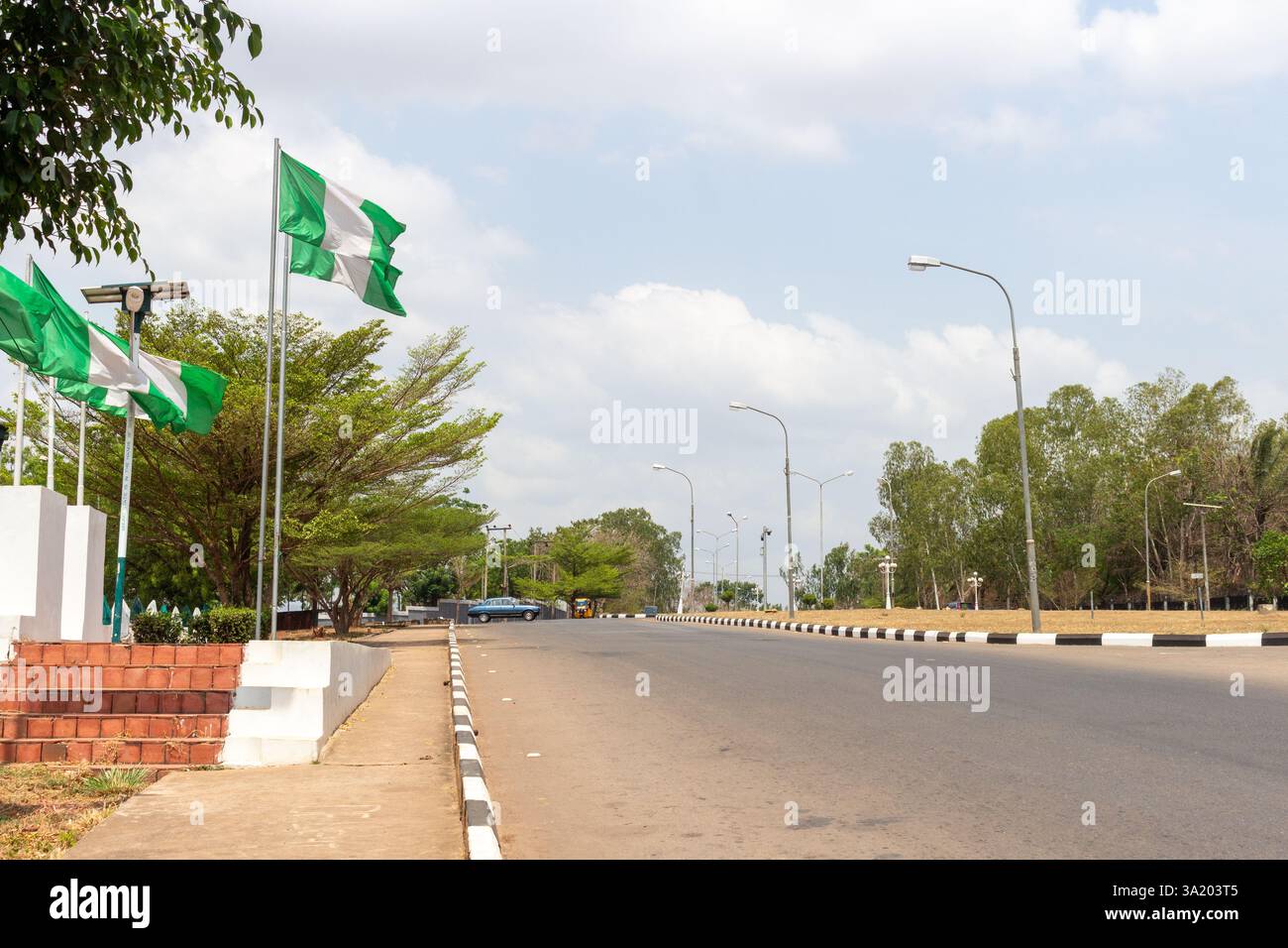 Una strada pulita e ben asfaltata a Enugu, Nigeria, fiancheggiata da bandiere nigeriane e lampioni sotto un cielo parzialmente nuvoloso. Marzo 2025 Foto Stock