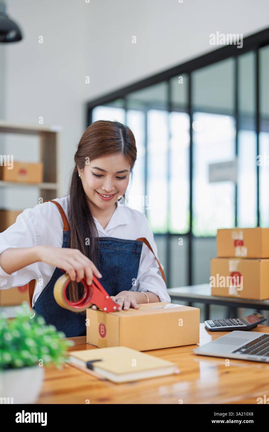 Una giovane donna è felice di confezionare le scatole con nastro adesivo in un moderno spazio di lavoro in casa, circondato da materiali di spedizione. Foto Stock
