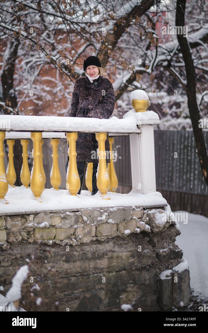 Una donna con un cappotto di pelliccia di karakul si trova accanto alla ringhiera di una casa storica, circondata dal fascino di una scena invernale innevata. Foto Stock