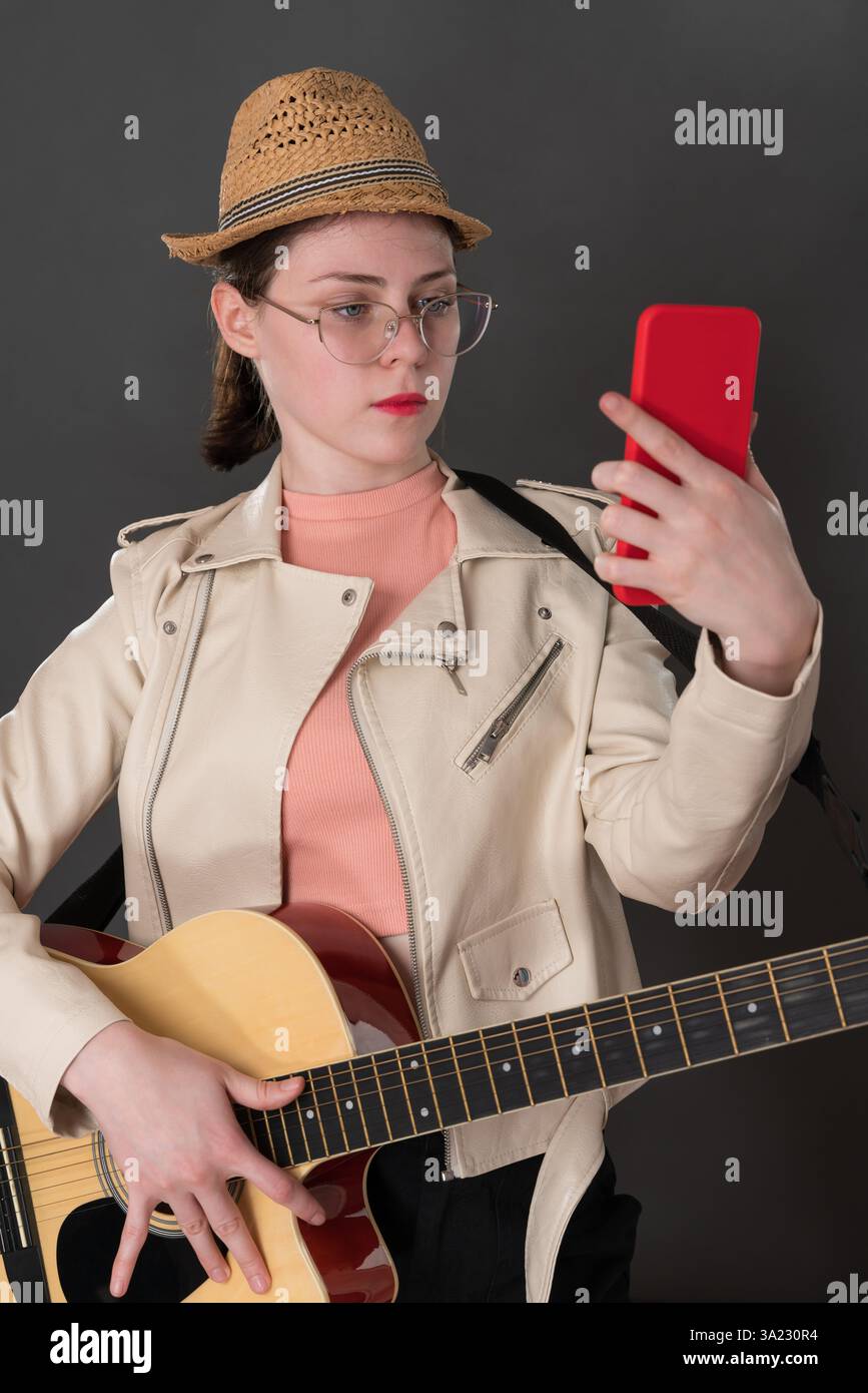 Giovane chitarrista con la chitarra che usa il telefono cellulare, che guarda un dispositivo di informazione portatile mentre impara a suonare la chitarra acustica a sei corde. Moderna Foto Stock