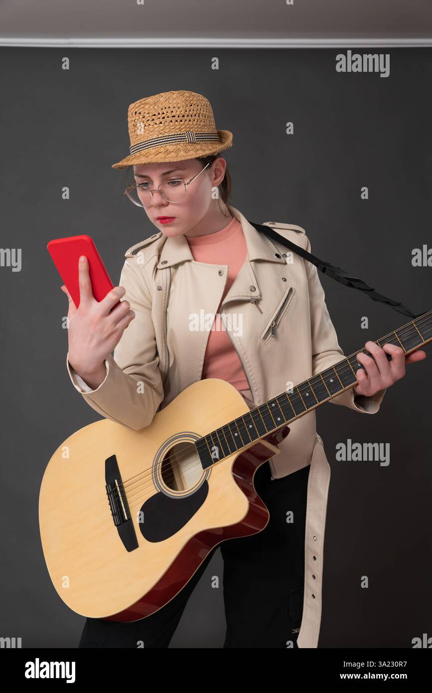 Chitarrista femminile con la chitarra che usa il telefono cellulare, osservandola imparando nuovi accordi, melodie o testi di canzoni mentre impara a suonare la chitarra. Musica e m Foto Stock