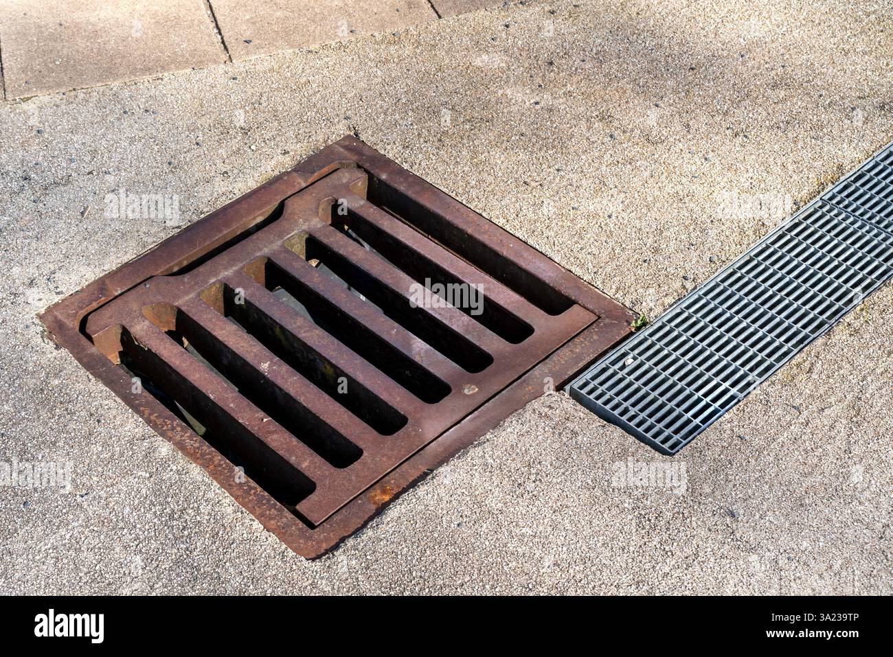 Primo piano di un vecchio coperchio di scarico in ghisa arrugginita accanto a un moderno canale di drenaggio in metallo incorporato in una superficie in cemento. Foto Stock