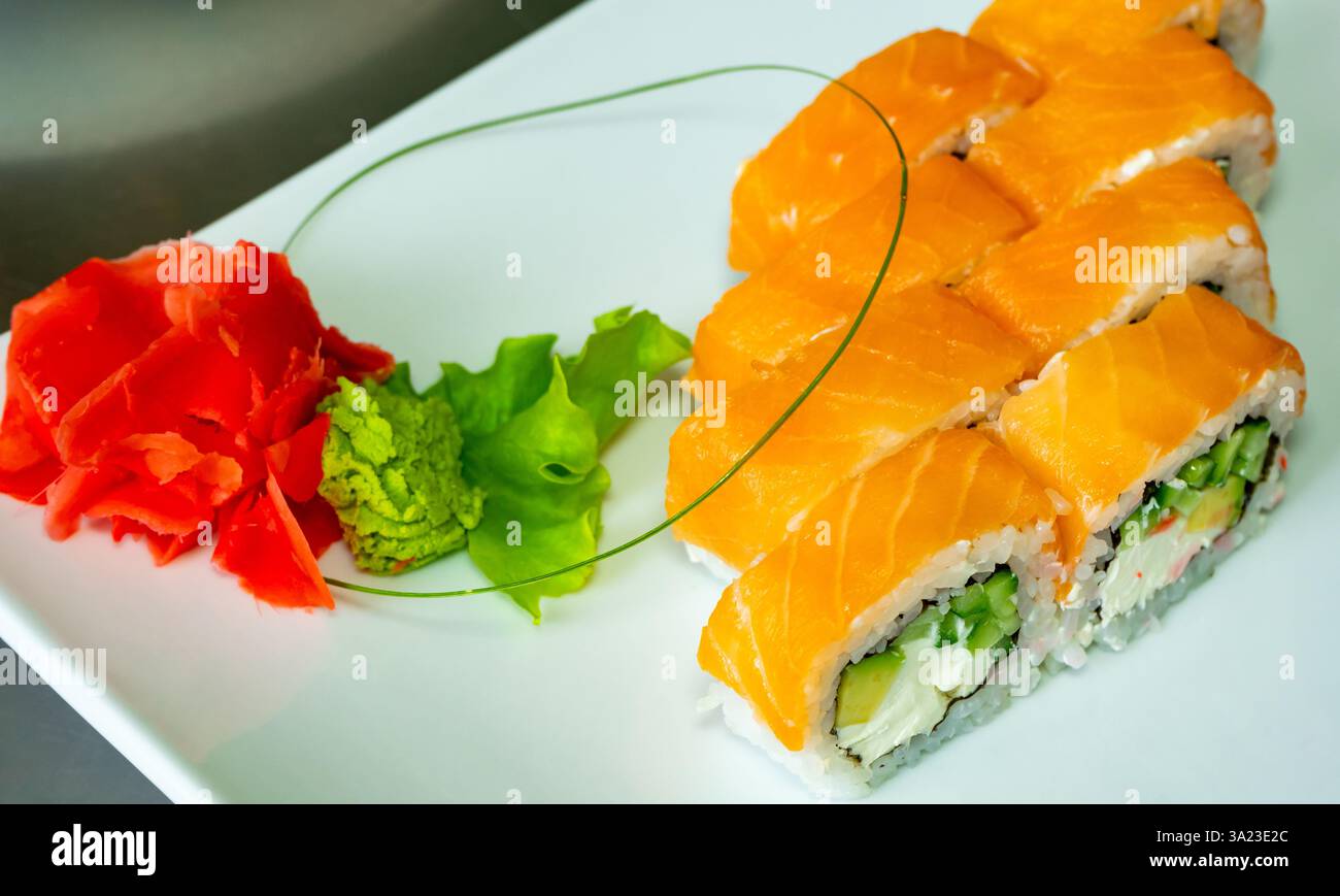 Gli involtini di sushi freschi sono disposti ordinatamente su un piatto bianco. Il piatto è caratterizzato da salmone vibrante, verdure croccanti ed è accompagnato da zenzero sott'aceto e. Foto Stock