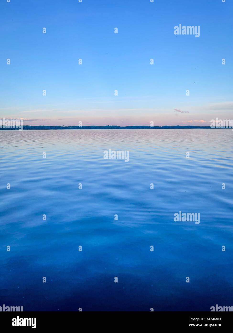 Un lago calmo si estende fino all'orizzonte, riflettendo un cielo azzurro limpido con morbide nuvole. La superficie dell'acqua presenta ondulazioni delicate. Foto Stock