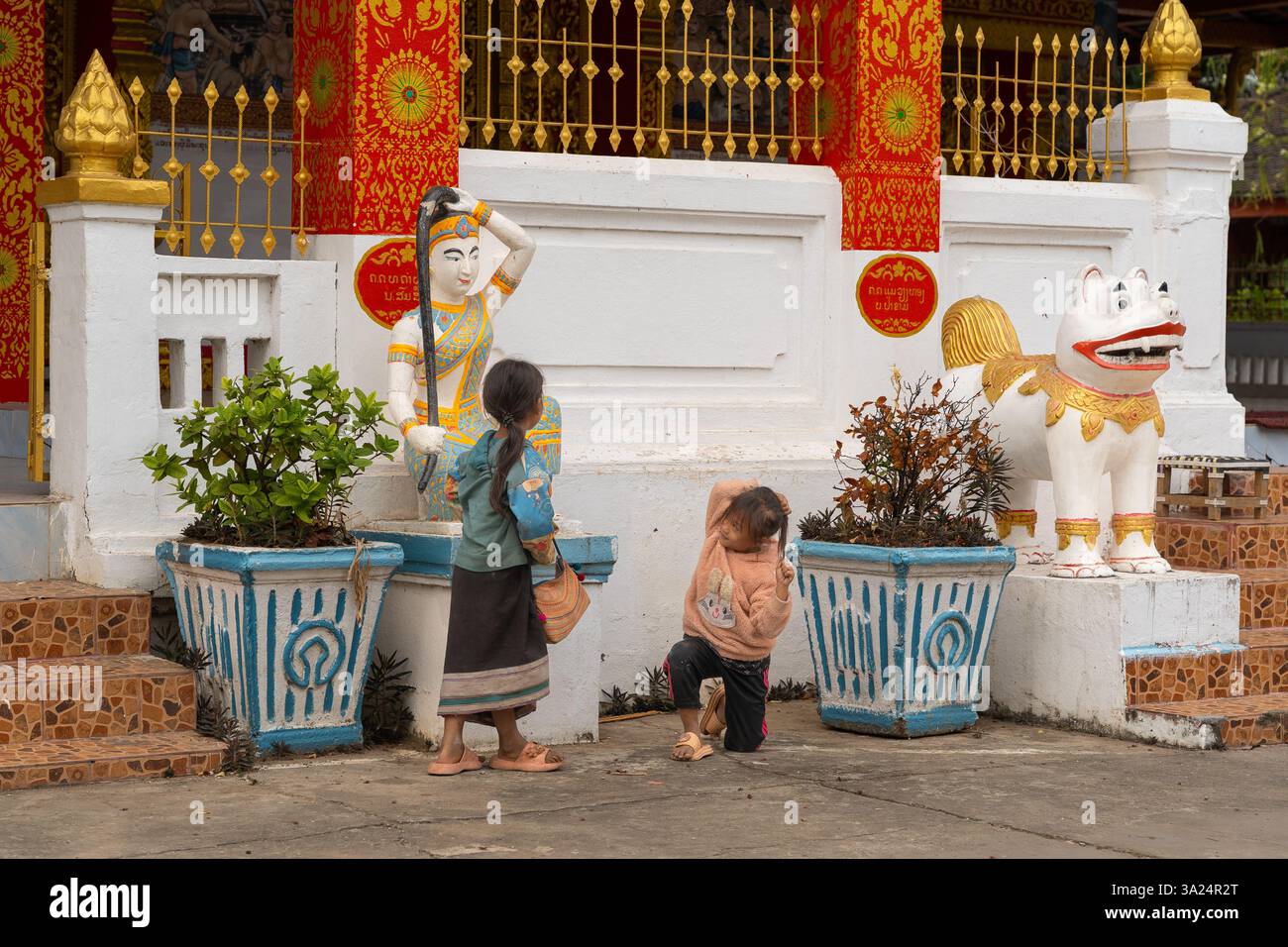 Bambini che giocano ad una scultura di cani a guardia della scala principale del tempio Phonsai Sanasongkham a Luang Prabang, Laos Foto Stock